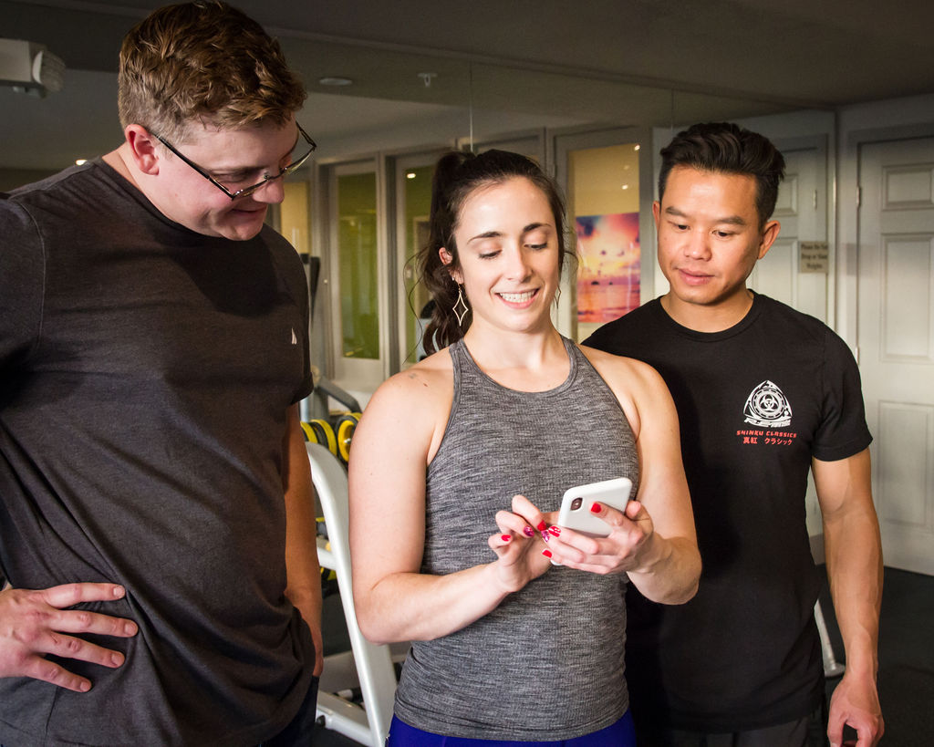 Two men in gym clothes smiling and looking at a Smartphone held by female fitness instructor