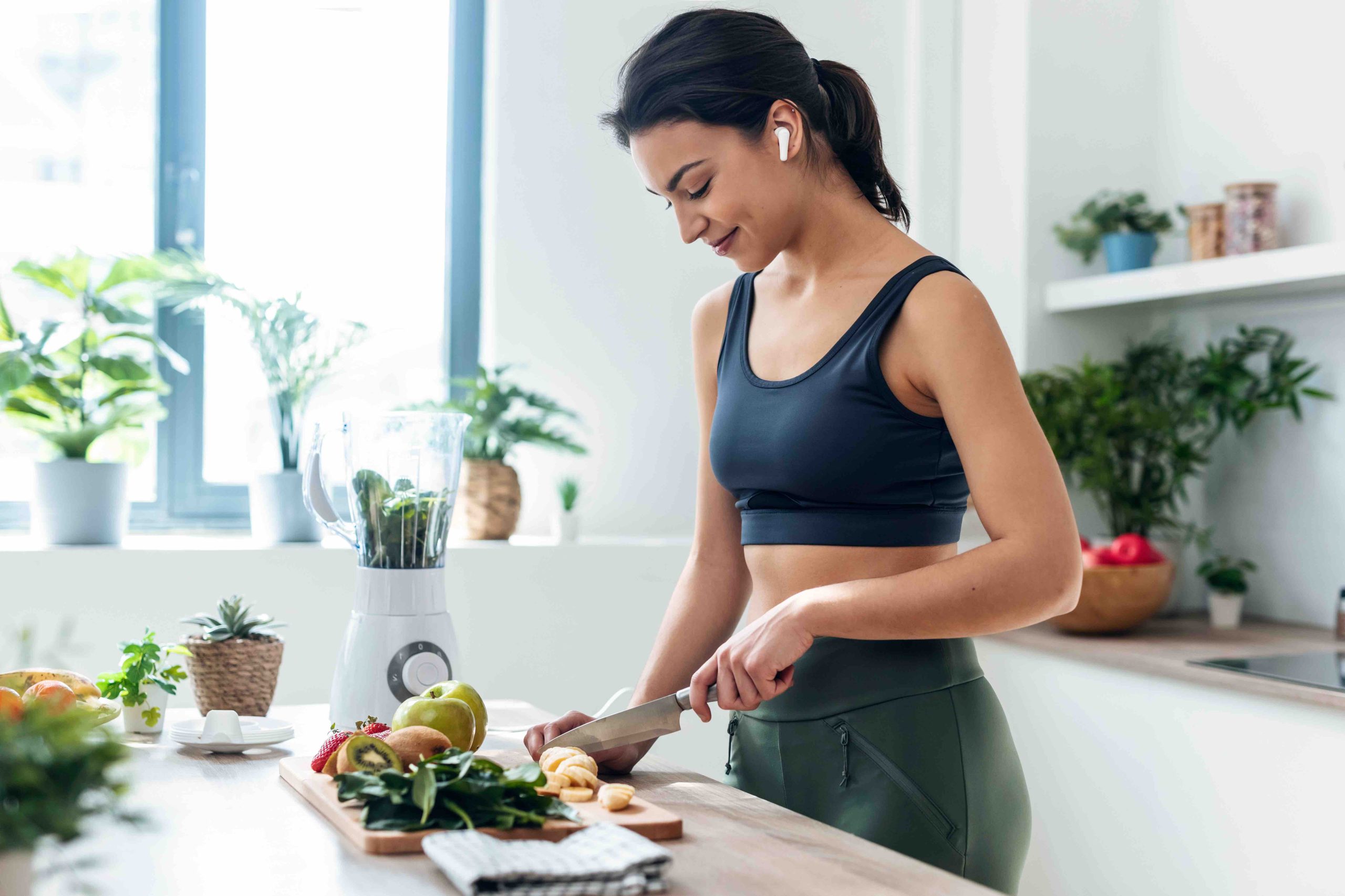 Shoot of athletic woman cutting fruits and vegetables to prepare a smoothie while listening to music with earphones in the kitchen at home